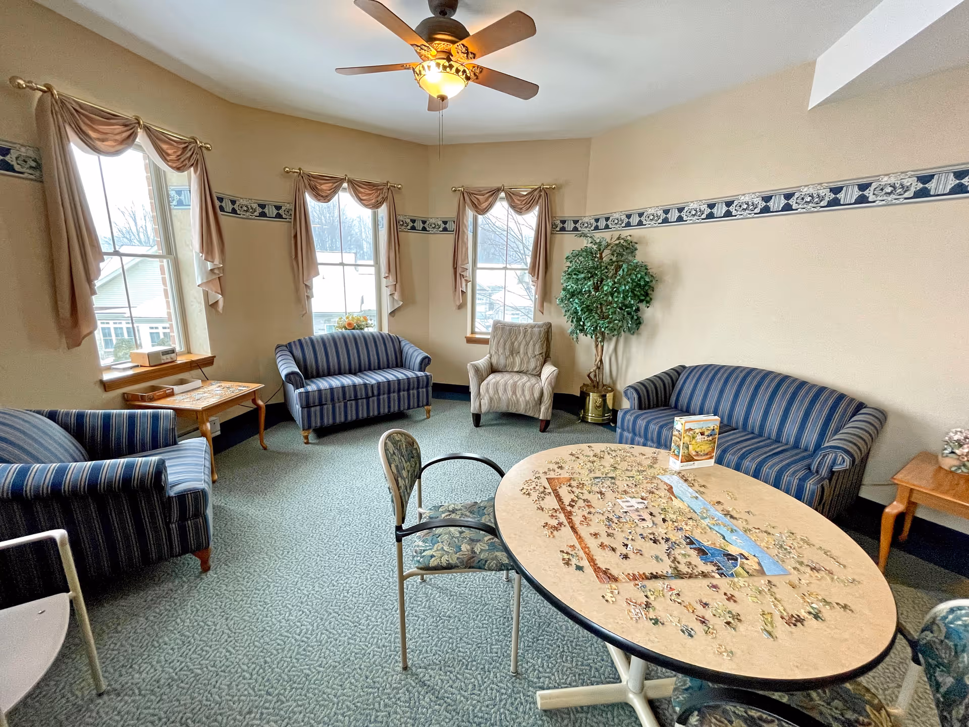 Cozy common room with striped sofas and armchairs around a table where a jigsaw puzzle is being assembled, with windows, drapes, and a ceiling fan.