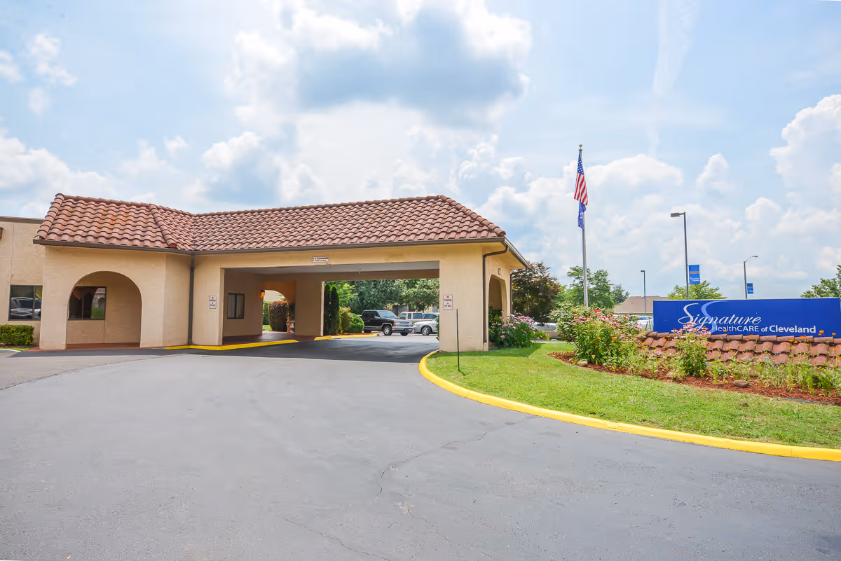 Exterior view of Signature HealthCARE of Cleveland showing the entrance driveway with a covered drop-off area, a landscaped garden with flowers, an American flag, and a facility sign on the right side.