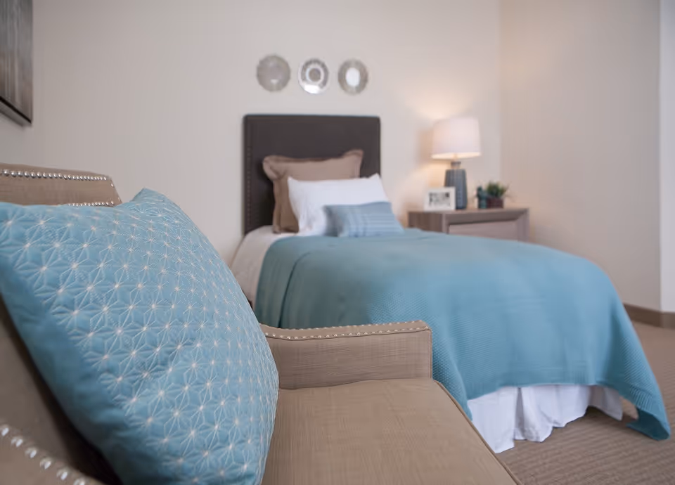 A cozy bedroom featuring a single bed with a dark upholstered headboard, layered pillows in white, brown, and blue, and a blue blanket. Next to the bed is a wooden nightstand with a lamp, a framed photo, and a small plant. In the foreground, there is a beige armchair with a blue patterned cushion. The walls are light-colored with three decorative round mirrors above the bed.