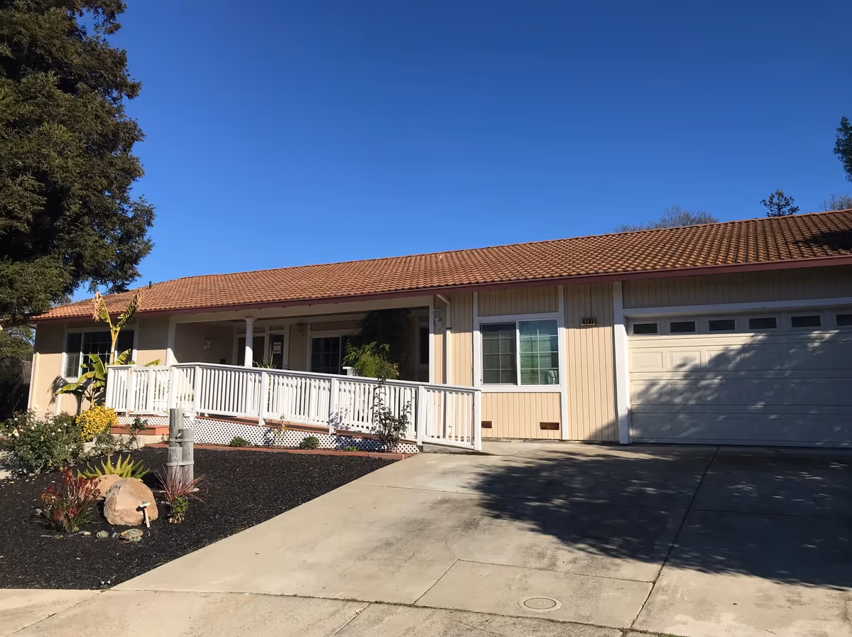 Single-story residential building with a beige exterior, a brown tiled roof, a white ramp leading to the front door, and a two-car garage. There are plants and small shrubs in the landscaped area near the driveway under a clear blue sky.