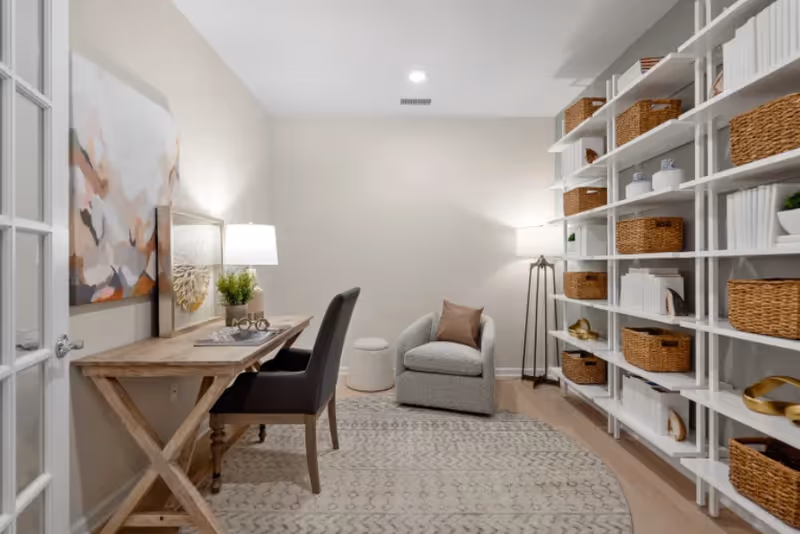 A cozy interior room featuring a wooden desk with a black chair, a small armchair with a brown pillow, a floor lamp, and a large white shelving unit filled with wicker baskets and decorative items. The room has light-colored walls, a patterned rug, and soft lighting.