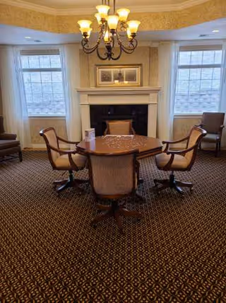 A cozy common room with a round table and chairs centered in front of a fireplace beneath a chandelier.