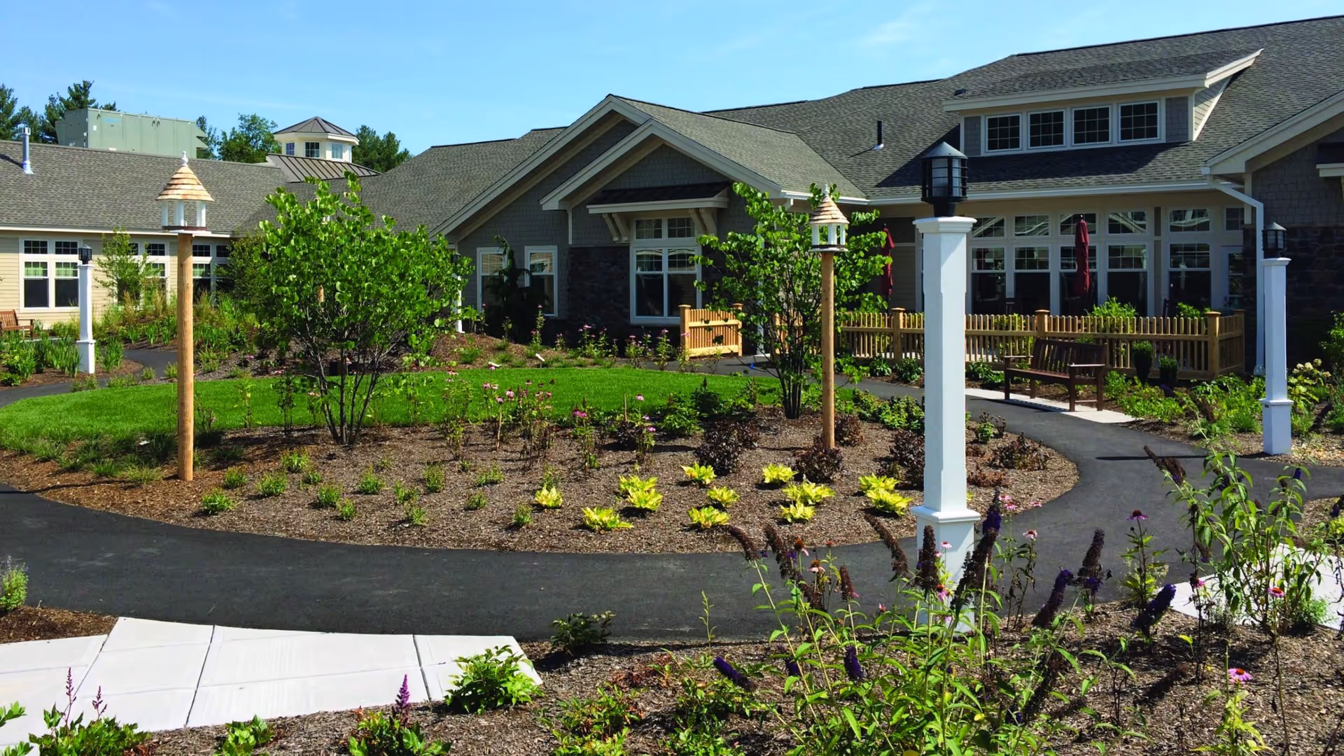 Outdoor garden area with paved walking paths, small trees, flower beds, birdhouses on wooden poles, and benches in front of a single-story building under a clear blue sky.