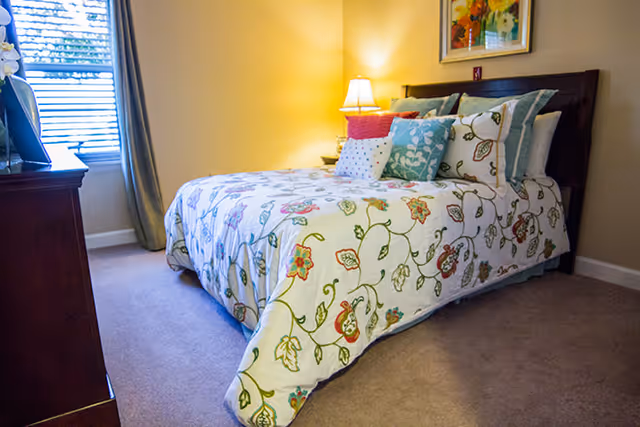A cozy bedroom with a bed covered in a floral patterned comforter and multiple decorative pillows. There is a wooden dresser on the left side near a window with blinds and curtains. A bedside table with a lamp is next to the bed, and a framed artwork hangs on the wall above the headboard.