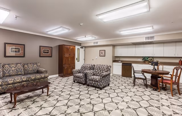 A spacious common area with patterned carpet flooring, featuring a floral-patterned sofa, two matching armchairs, a wooden coffee table, a round wooden dining table with four chairs, and a kitchenette with white cabinets and a countertop. The walls are beige with framed artwork, and the ceiling has fluorescent lighting.