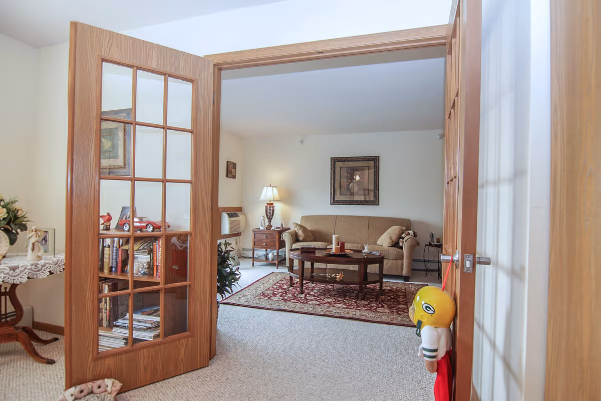 View through wooden French doors into a cozy living room with a beige sofa, a wooden coffee table on a red patterned rug, a side table with a lamp, framed artwork on the wall, and a small bookshelf with books and decorative items.