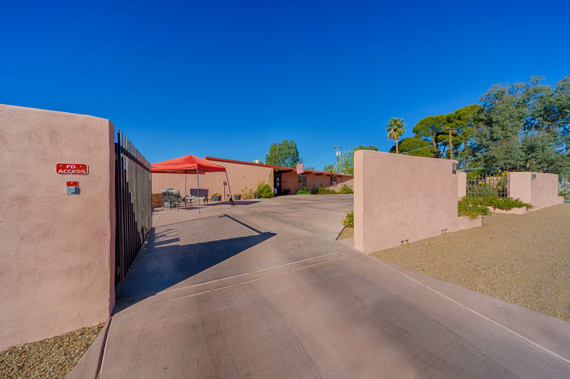 Entrance driveway to a facility with pink stucco walls and a black metal gate. There is a red canopy with outdoor seating on the left side near the building. The sky is clear and blue with some trees in the background.
