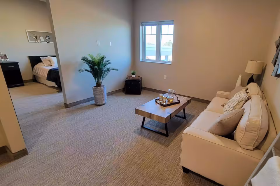 A cozy living room area with a beige sofa adorned with cushions, a wooden coffee table with a tray holding a pitcher and glasses, a floor lamp, and a potted plant near the wall. There is a window letting in natural light and an adjacent bedroom visible through an open doorway.