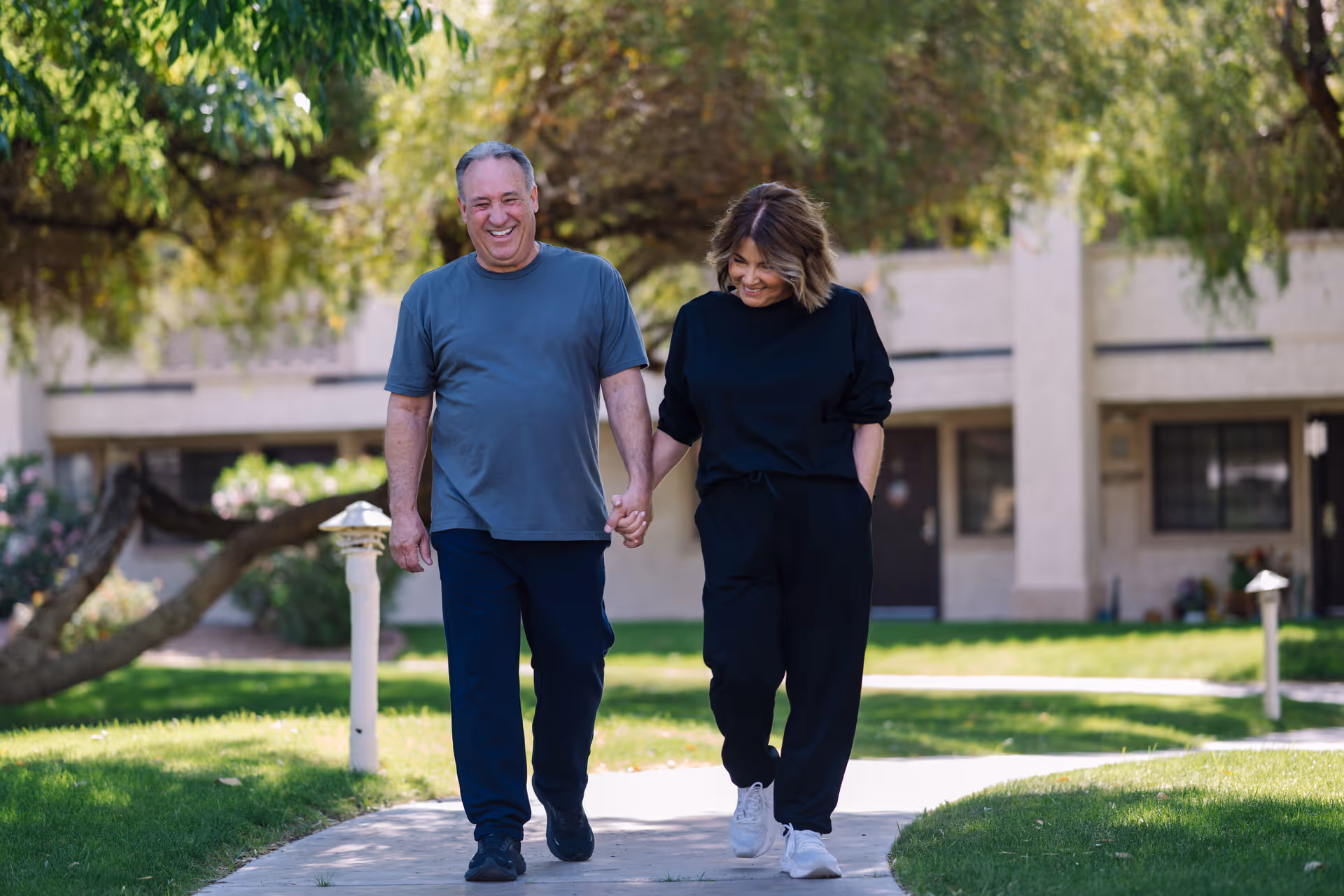 An older man and woman holding hands and walking on a sidewalk in a green outdoor area with trees and a building in the background, both smiling and dressed casually.