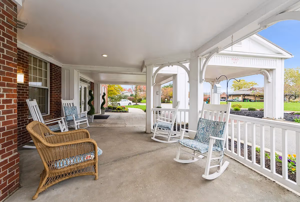 Covered front porch with white rocking chairs, a wicker chair, and views of a landscaped lawn and driveway.