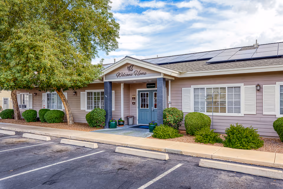 Front entrance of a single-story senior living building with a 'Welcome Home' sign, shrubs, and a parking area.