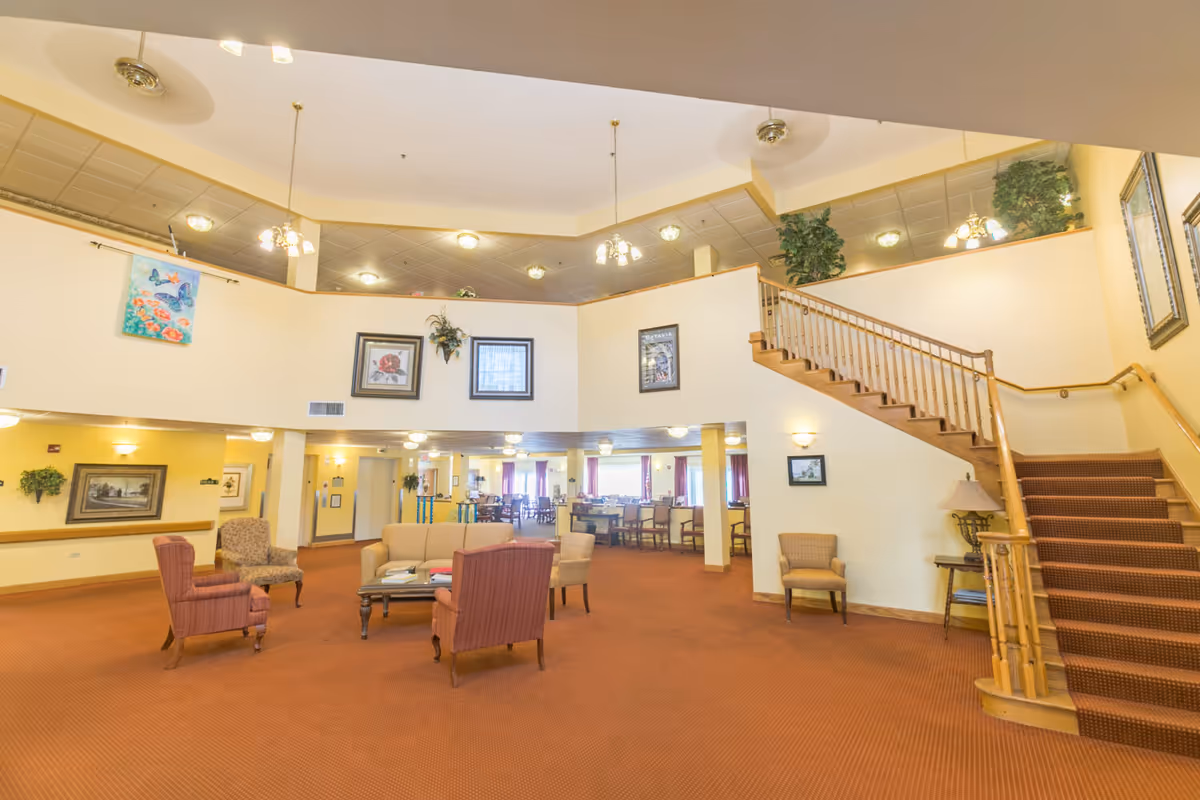 Spacious senior living facility lobby with seating, a central sitting area, and a staircase leading to an upper level and dining area.