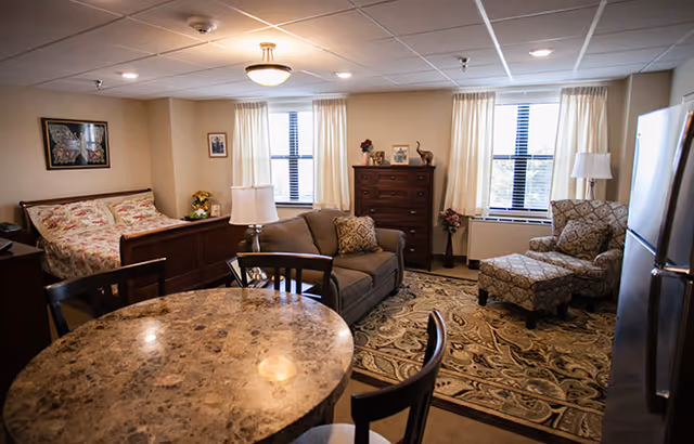 A cozy senior living studio apartment featuring a bed with floral bedding, a small sofa with a decorative pillow, an armchair with an ottoman, a wooden dresser, a round dining table with chairs, and a refrigerator. The room has three windows with cream-colored curtains and a patterned area rug on the floor.
