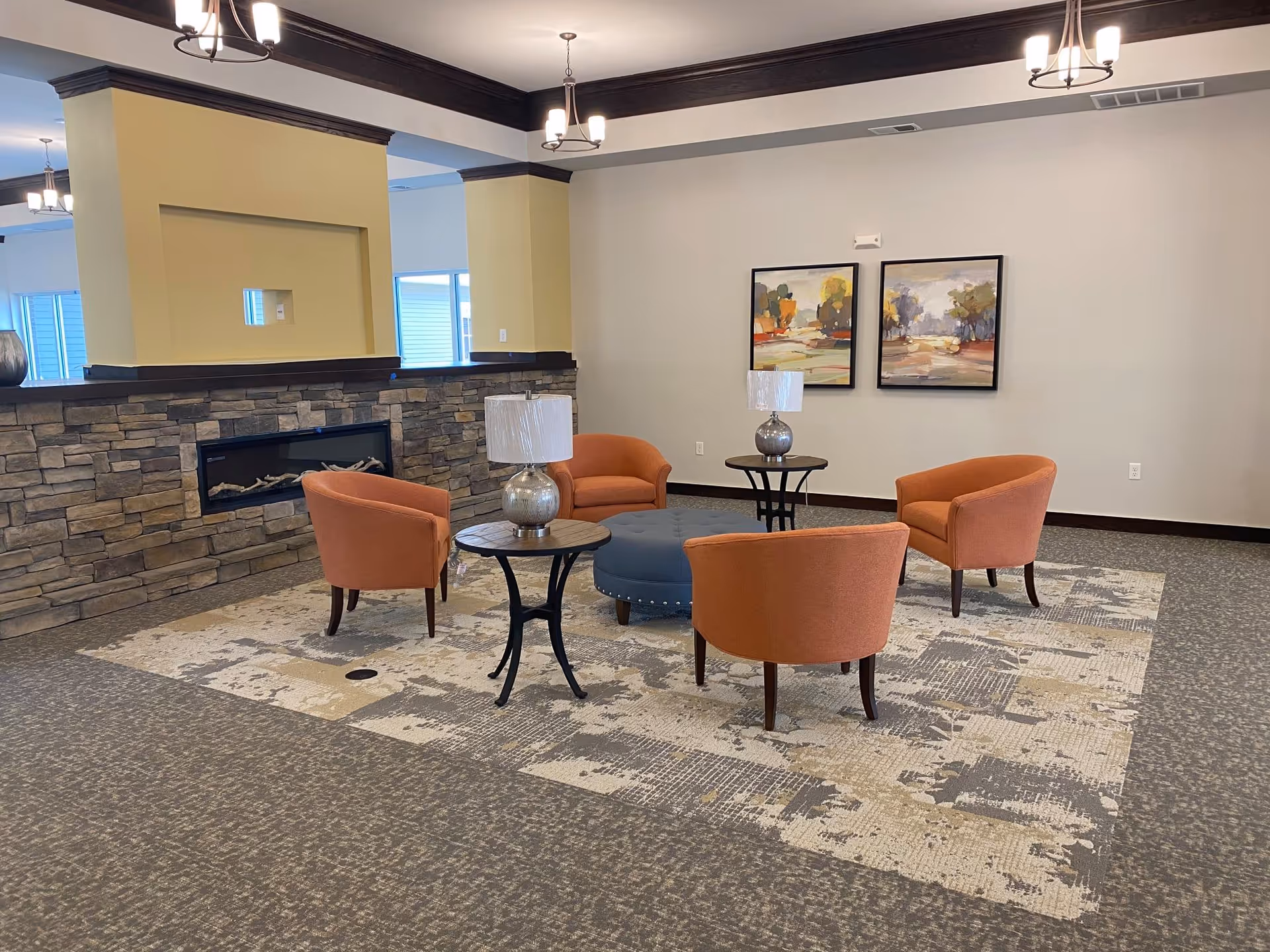 A cozy seating area in a senior living facility with four orange armchairs arranged around a round blue ottoman. Two small round tables with silver lamps are placed between the chairs. The room features a stone fireplace with a yellow mantle, neutral-colored walls, two framed landscape paintings, and modern ceiling light fixtures.