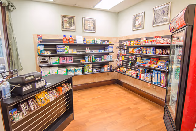 Small convenience store area inside a facility with shelves stocked with snacks, beverages, household items, and personal care products. There is a counter with a cash register and a refrigerated display on the right side. The room has light-colored walls, wooden flooring, and framed artwork on the walls.