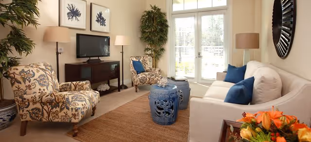 Bright living room with a white sofa, patterned armchairs, a TV console, blue decorative stools used as coffee tables, potted plants, and French doors letting in daylight.