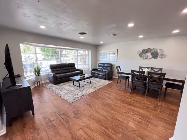 A bright living room and dining area with wooden flooring. The living room features two black leather sofas, a coffee table on a patterned rug, and a TV on a stand near a large window with blinds. The dining area has a dark wooden table with six chairs and a decorative wall piece above it.