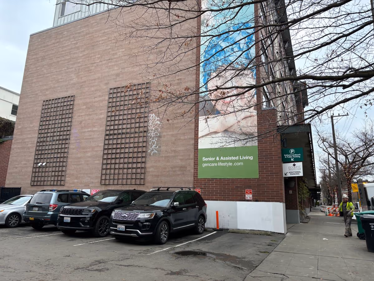 Exterior view of a brick senior living building with a large vertical banner reading 'Senior & Assisted Living' above a small parking lot with parked cars and a sidewalk.
