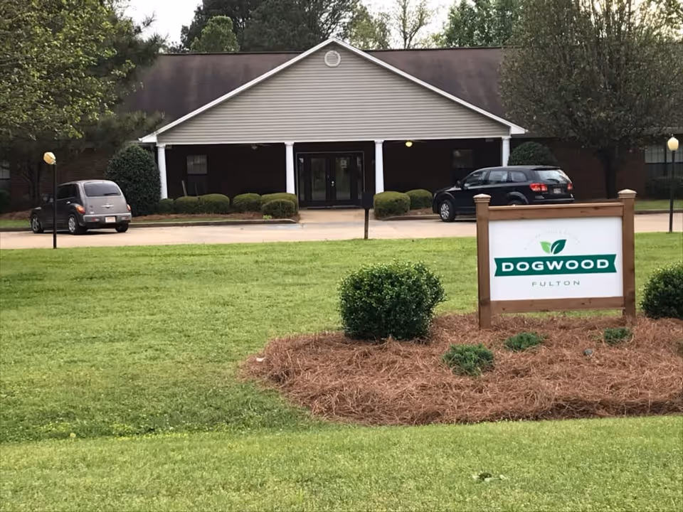 Front exterior view of a single-story building with a peaked roof and a covered entrance supported by white columns. There are two cars parked in front of the building, surrounded by neatly trimmed bushes and trees. In the foreground, there is a landscaped area with mulch and shrubs, and a wooden sign that reads 'DOGWOOD FULTON' with a green leaf logo above the text.