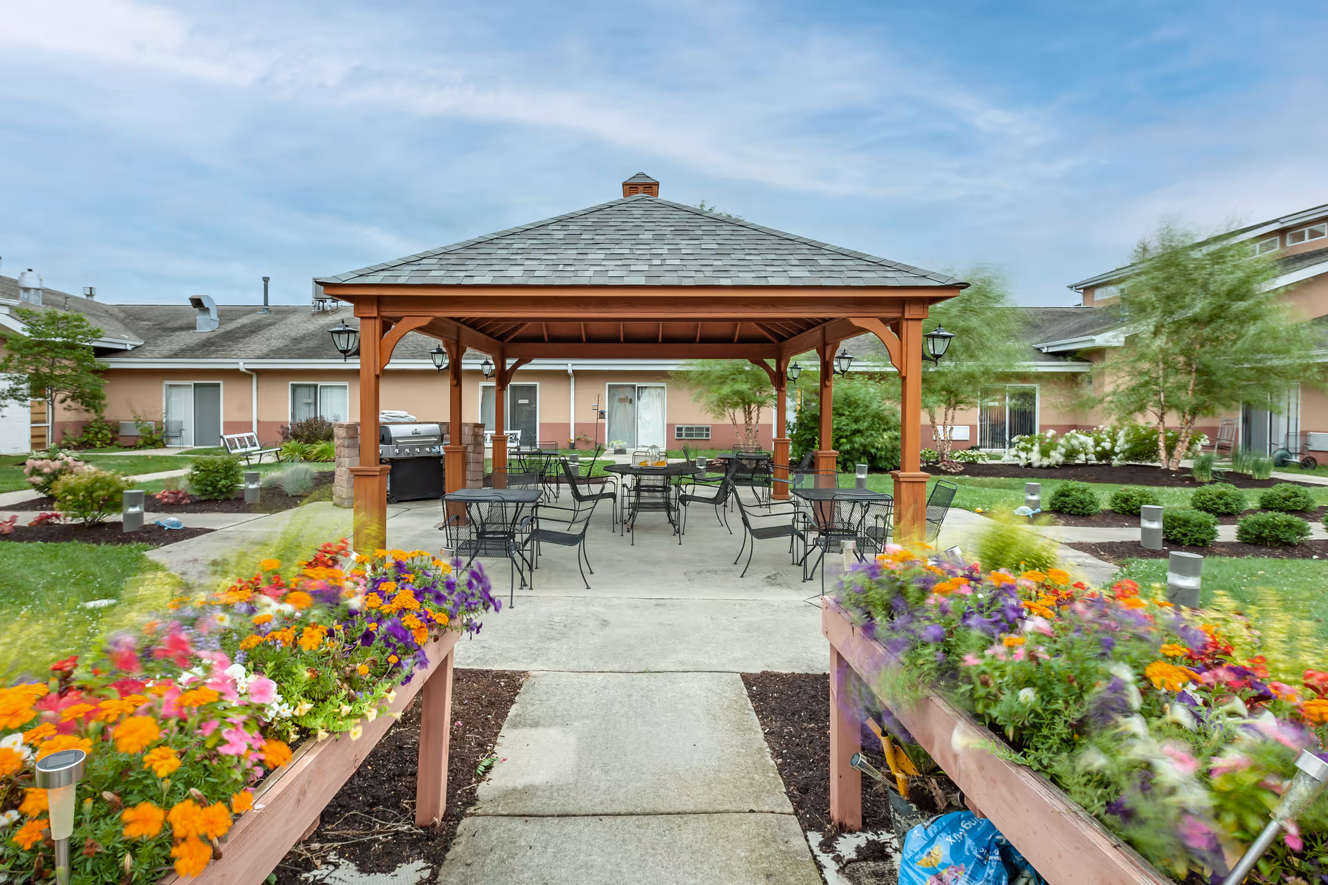 Outdoor seating area with a wooden gazebo covering several black metal tables and chairs, surrounded by colorful flower beds and greenery, with a building in the background under a partly cloudy sky.