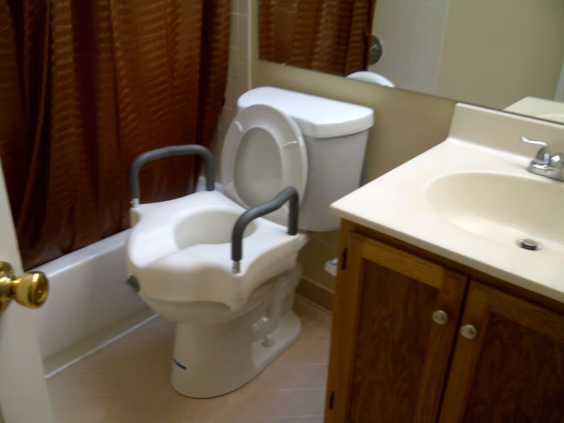 Small bathroom with a raised toilet seat with arm supports next to a sink and vanity.