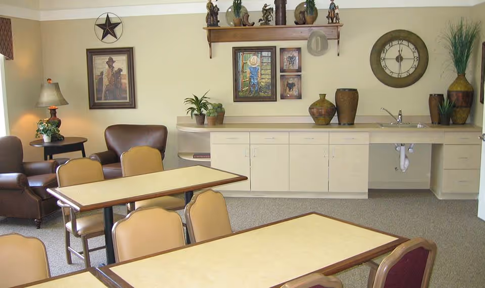 A cozy common area with beige walls featuring framed western-themed artwork and a large round clock. The room contains several tables with beige tops and chairs arranged around them. In the background, there is a countertop with cabinets underneath, decorated with various vases and potted plants. Two brown leather armchairs and a small side table with a lamp and plant are positioned near the wall.