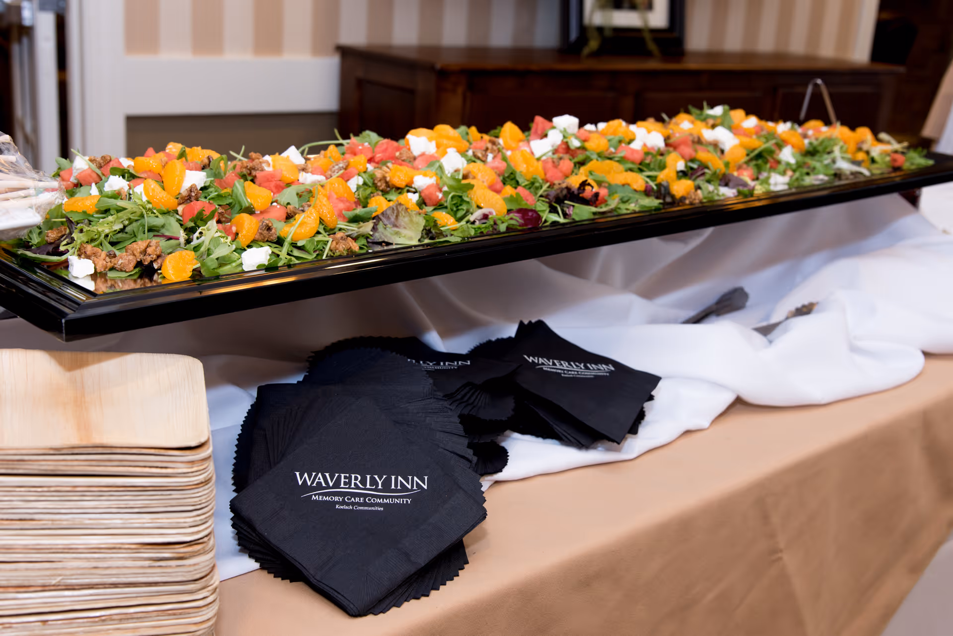 A large rectangular tray filled with a colorful salad containing mixed greens, mandarin orange slices, grapefruit pieces, walnuts, and chunks of white cheese, placed on a table with a beige tablecloth and white fabric underneath. Next to the tray are stacks of wooden plates and black napkins with the text 'Waverly Inn Memory Care Community' printed on them.