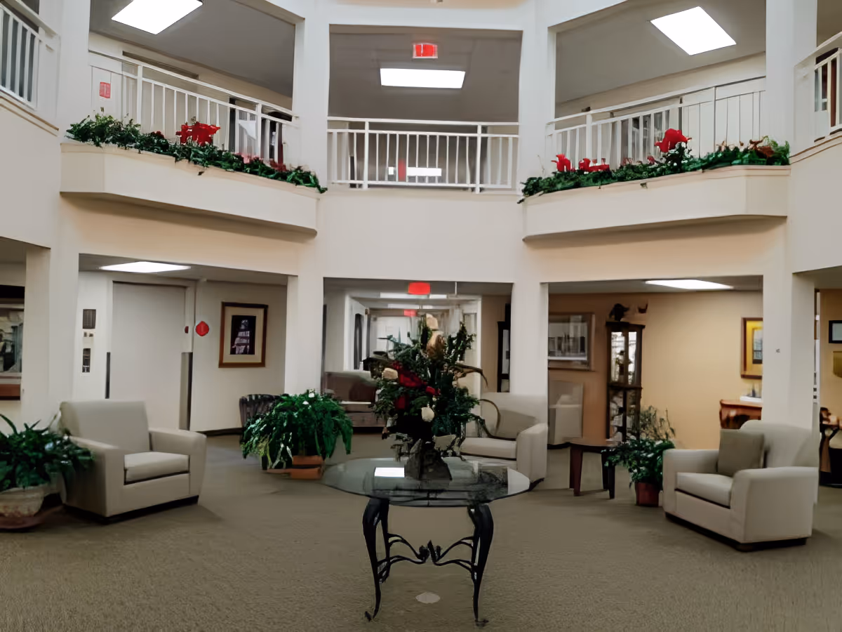 Atrium-style common lounge with armchairs, potted plants, a glass-top table with a floral arrangement, and a second-floor balcony.