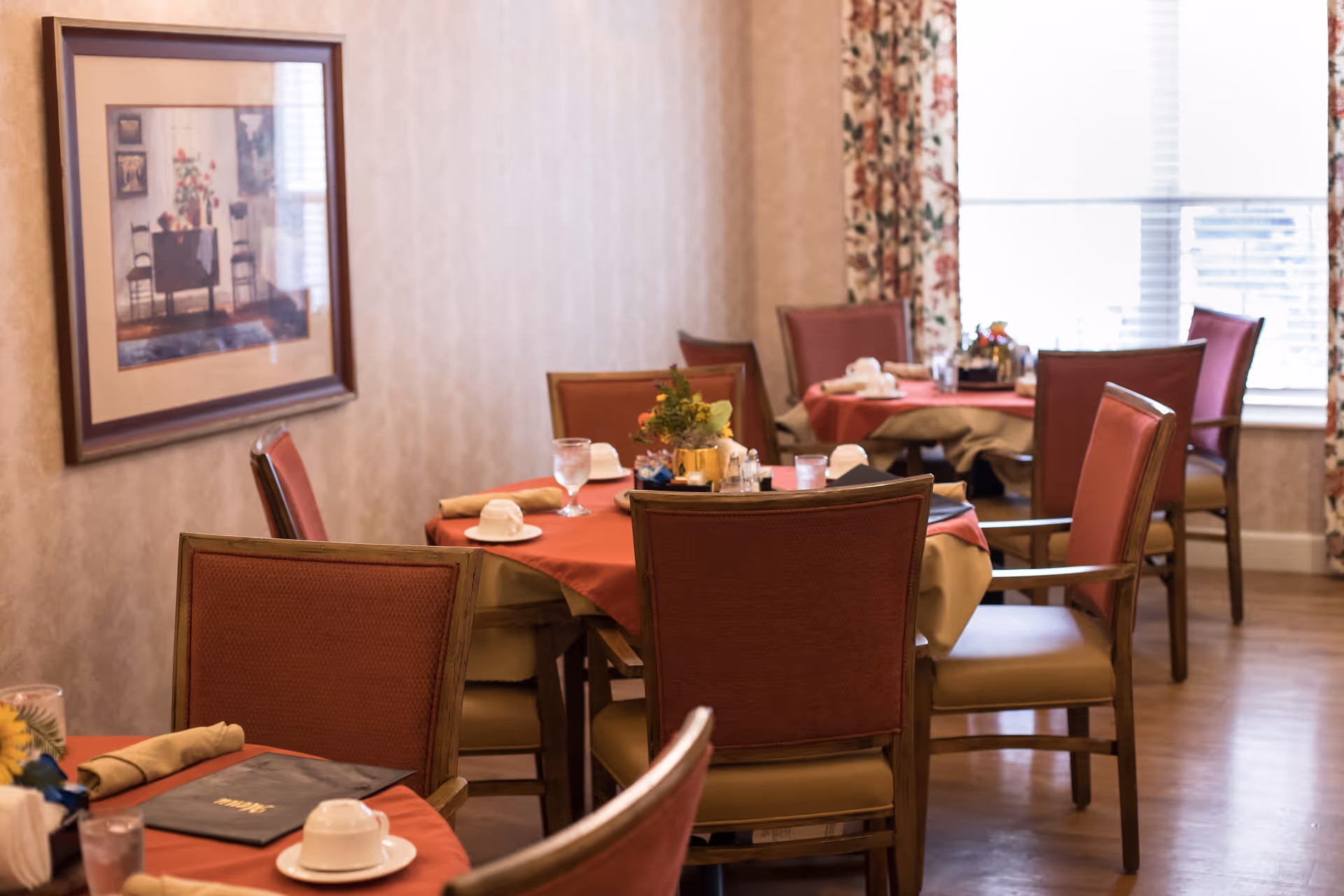 A cozy dining room with several tables covered in red and beige tablecloths. Each table is set with cups, glasses, napkins, and small floral centerpieces. The room has wooden chairs with red upholstery and a large window with floral curtains allowing natural light to enter. A framed picture hangs on the wall.