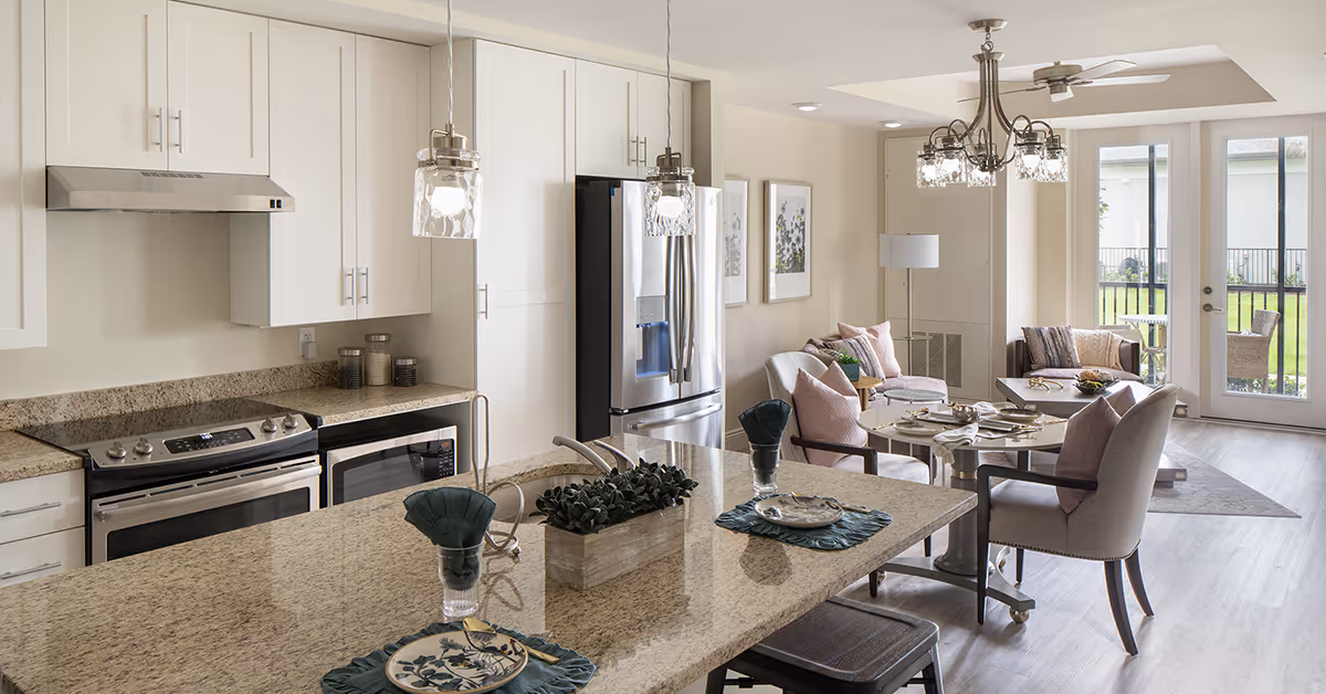 A modern open-concept kitchen and living area with granite countertops, stainless steel appliances, and a dining table set for four. The living area has comfortable chairs with pink cushions, a coffee table, and large windows with a door leading outside.