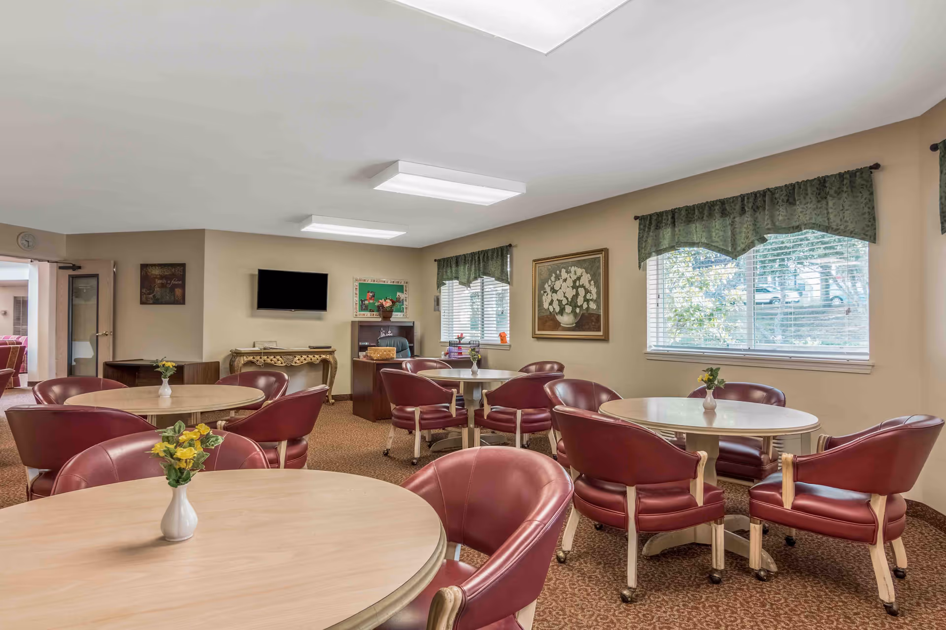A well-lit common room with several round tables surrounded by red cushioned chairs on wheels. Each table has a small white vase with yellow flowers. The room has beige walls, green valance curtains on the windows, a wall-mounted TV, a decorative console table, and framed artwork. There is a carpeted floor with a patterned design.