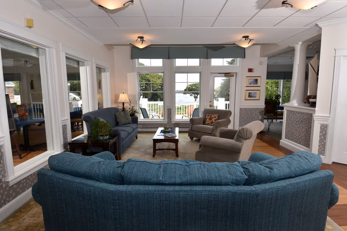 A cozy living room area with a blue sofa in the foreground, two armchairs, a coffee table, and a side table with a lamp. Large windows and a glass door provide a view of an outdoor patio and greenery beyond. The room has white walls, decorative molding, and a patterned carpet.