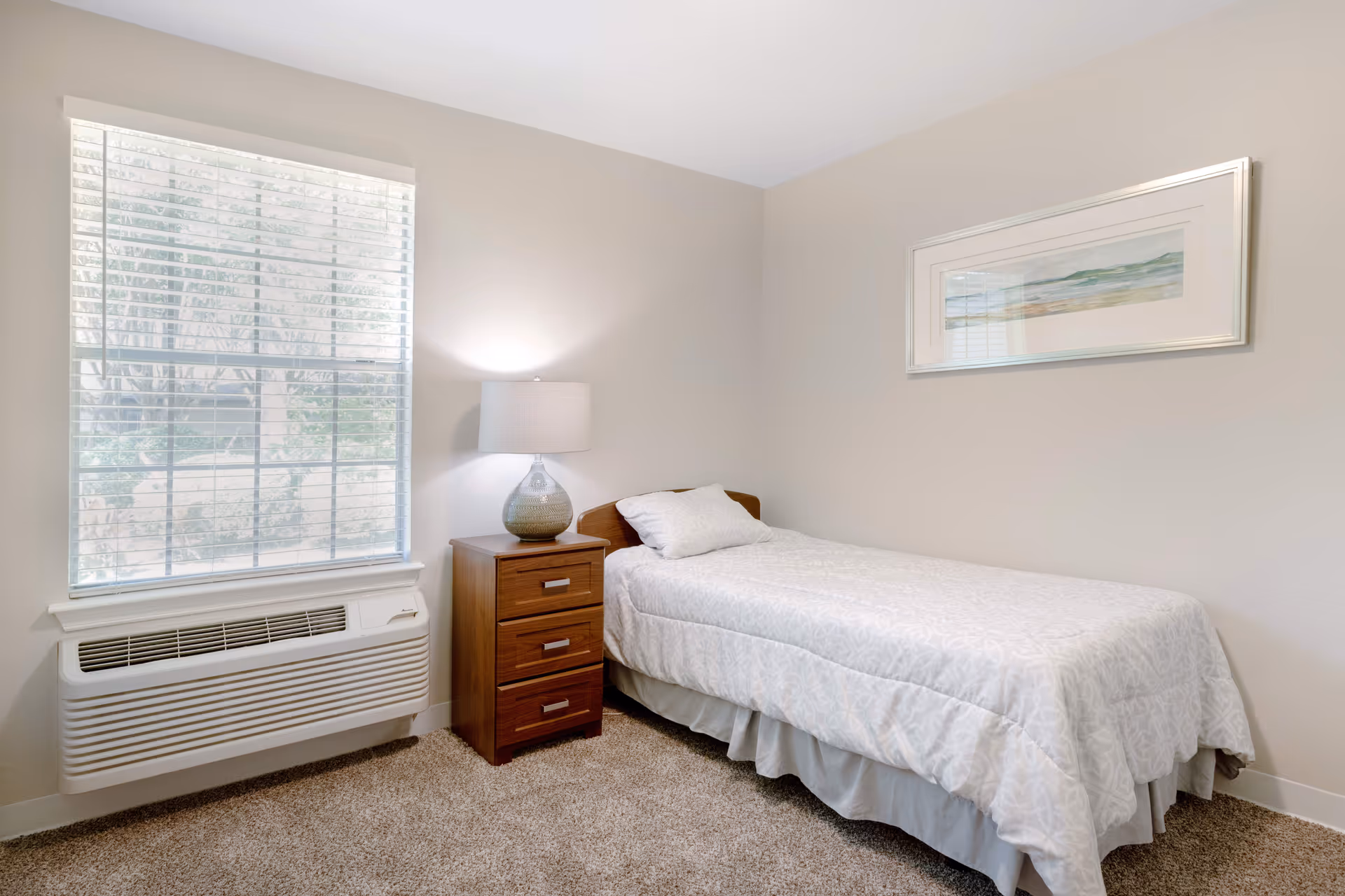 A small, neatly arranged bedroom with a single bed covered in a white patterned bedspread. Next to the bed is a wooden nightstand with three drawers and a decorative lamp on top. A large window with white blinds lets in natural light, and a framed landscape painting hangs on the beige wall above the bed. The floor is carpeted in a light brown color.