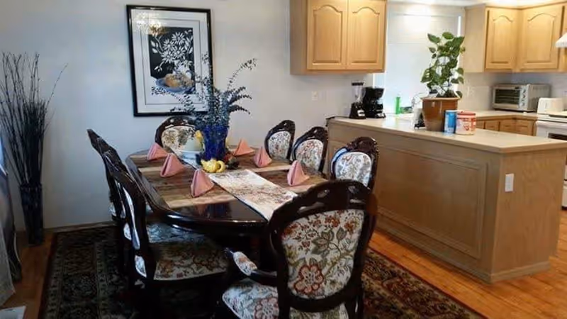 Ornate wooden dining table set with six upholstered chairs and place settings in an open kitchen-dining area.