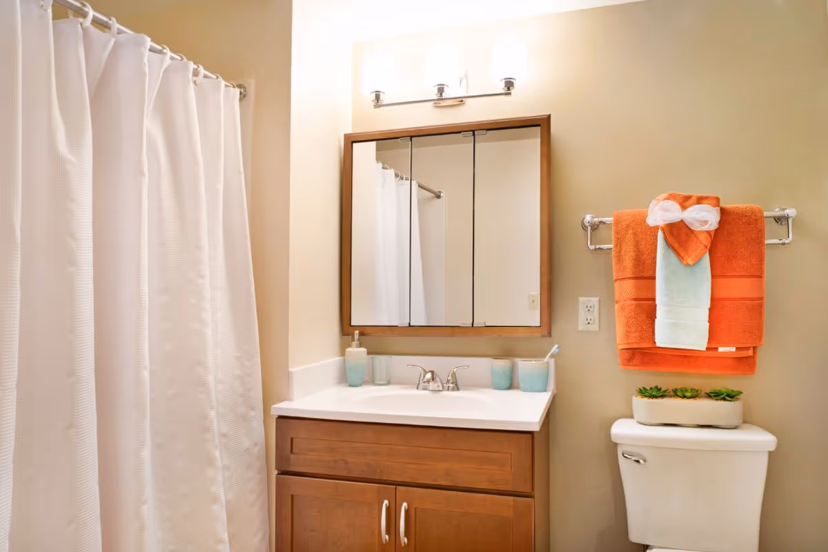 A clean bathroom with a white shower curtain on the left, a wooden vanity with a white countertop and a sink in the center, and a mirror with three panels above the sink. On the right, there is a white toilet with a small planter of succulents on top and a towel rack above it holding orange and light blue towels, with a decorative white bow on the orange towel.