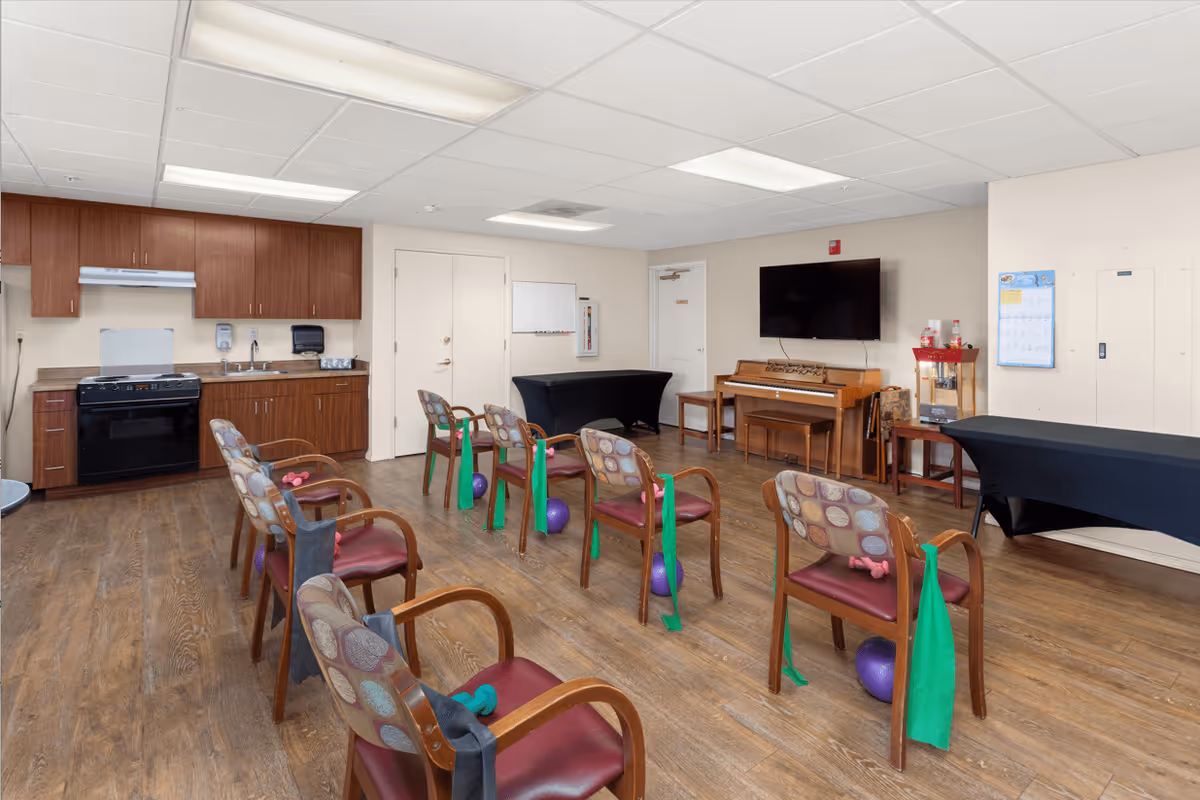 A multipurpose room with wooden flooring and a kitchenette on the left side. Several chairs are arranged in rows, each with exercise bands and small weights placed on or near them. A piano and a wall-mounted TV are visible on the right side, along with tables covered in black cloth. The room is well-lit with ceiling lights.