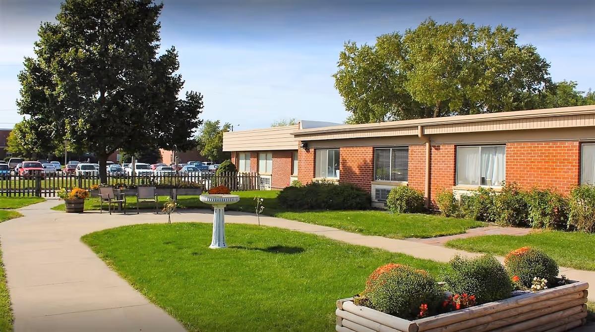 Outdoor courtyard area of a senior living facility with a paved walkway, green grass, a birdbath, benches, and flower beds. The building is a single-story brick structure with windows and surrounded by trees and a wooden fence.
