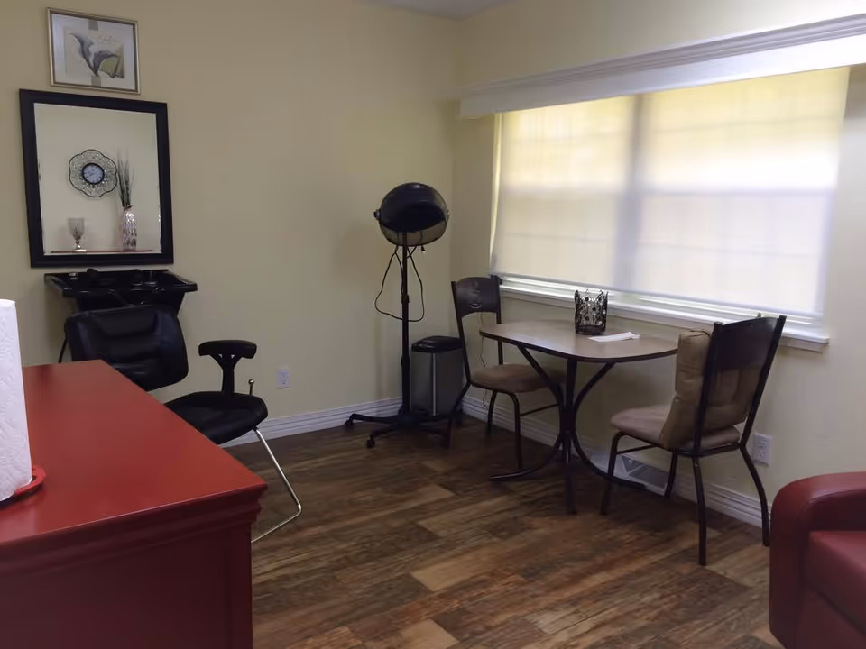 Small interior room with a salon chair and hood dryer, a shampoo sink, and a small table with two chairs by a window.