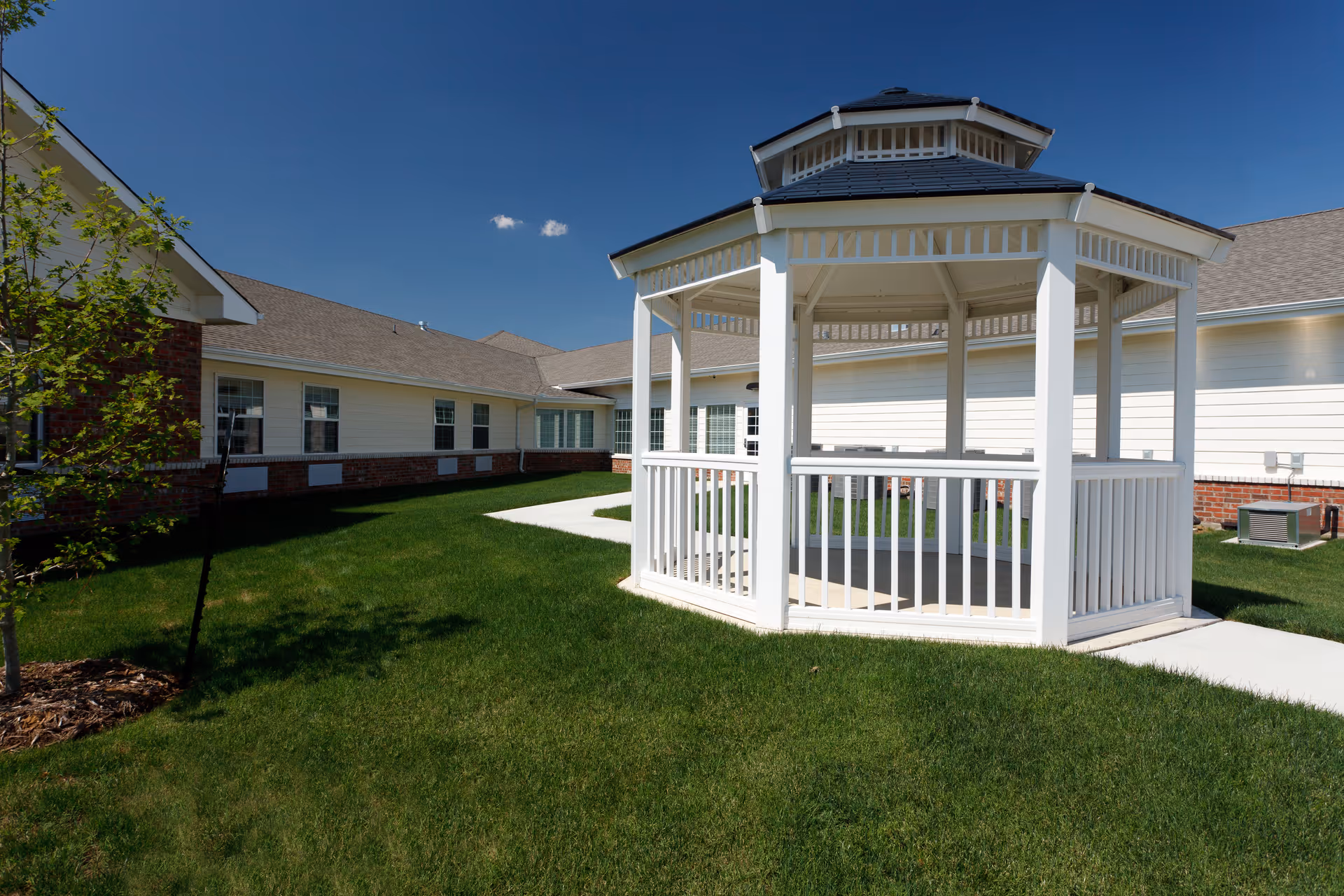 A white wooden gazebo with a dark roof situated on a well-maintained green lawn, surrounded by a single-story building with white siding and brick accents under a clear blue sky.