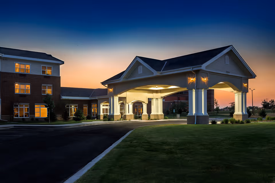 Front entrance porte-cochère of a brick retirement community building lit at dusk with a driveway and lawn.