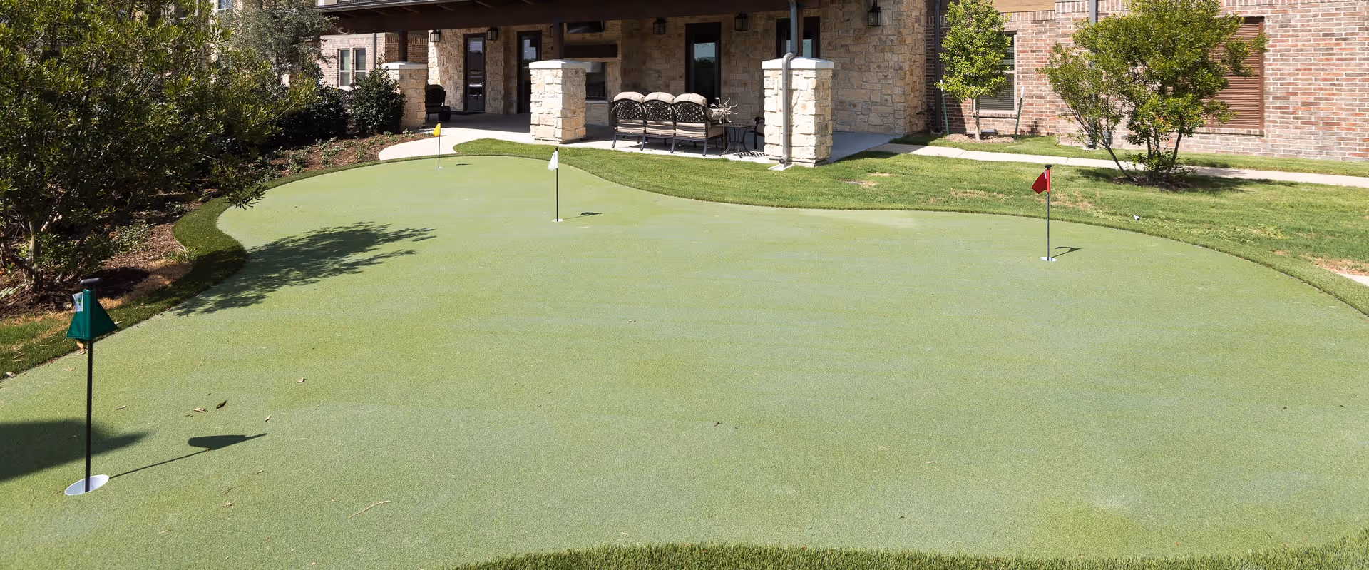 Outdoor putting green with three golf holes and flags in front of a brick building with stone pillars and a covered patio area with seating.