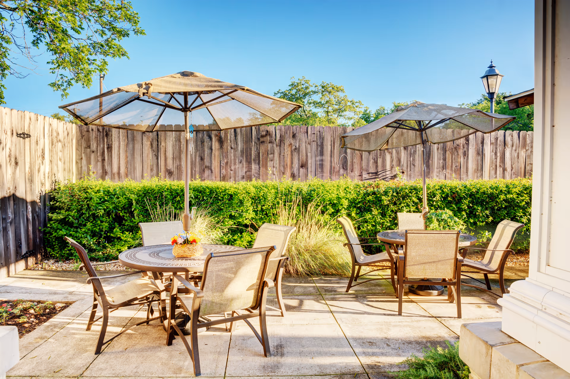 Outdoor patio area with two round tables, each surrounded by four chairs and shaded by large umbrellas. The patio is enclosed by a wooden fence with green bushes and plants along the fence line. A lamppost is visible in the background under a clear blue sky.