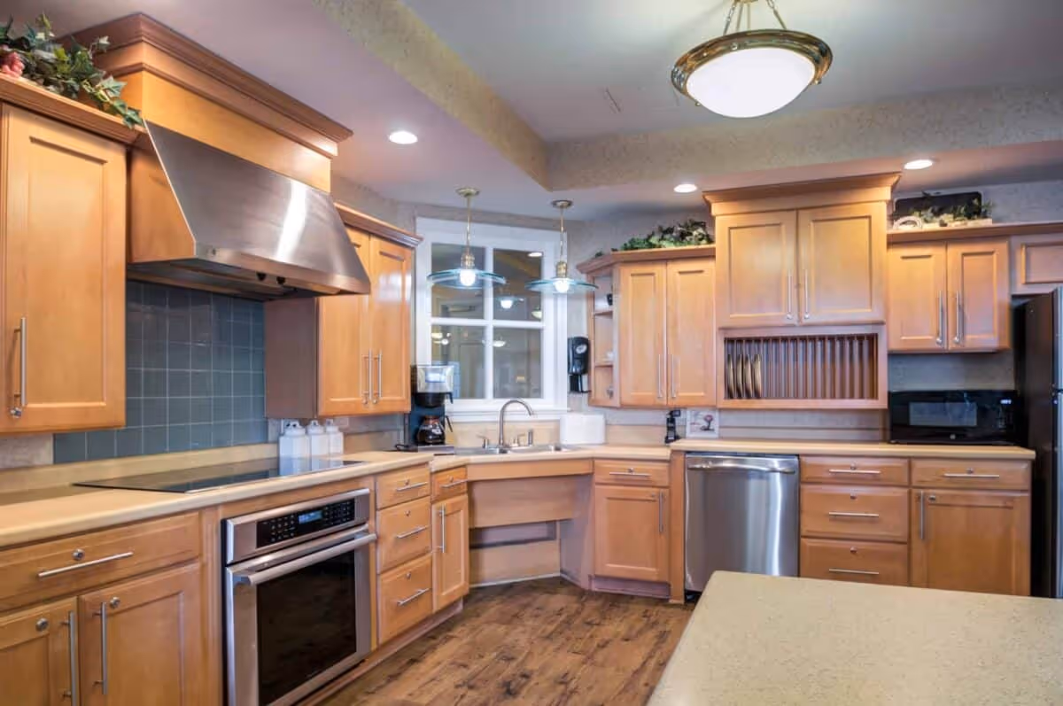A modern kitchen with wooden cabinets, stainless steel appliances including an oven, dishwasher, and refrigerator. The kitchen features a double sink under a window, pendant lights hanging from the ceiling, and a wooden floor. There are decorative plants on top of the cabinets.