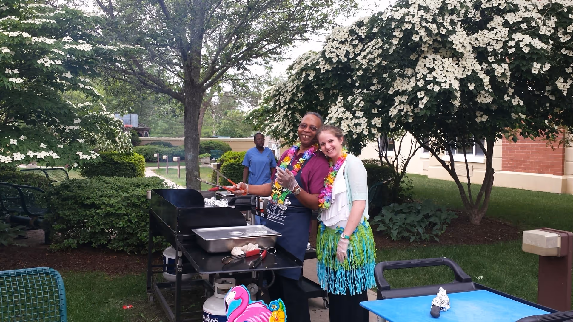 Two women smiling and posing together near a barbecue grill outdoors, both wearing colorful leis and grass skirts, with a third person in blue scrubs standing in the background near trees and bushes in a garden area.