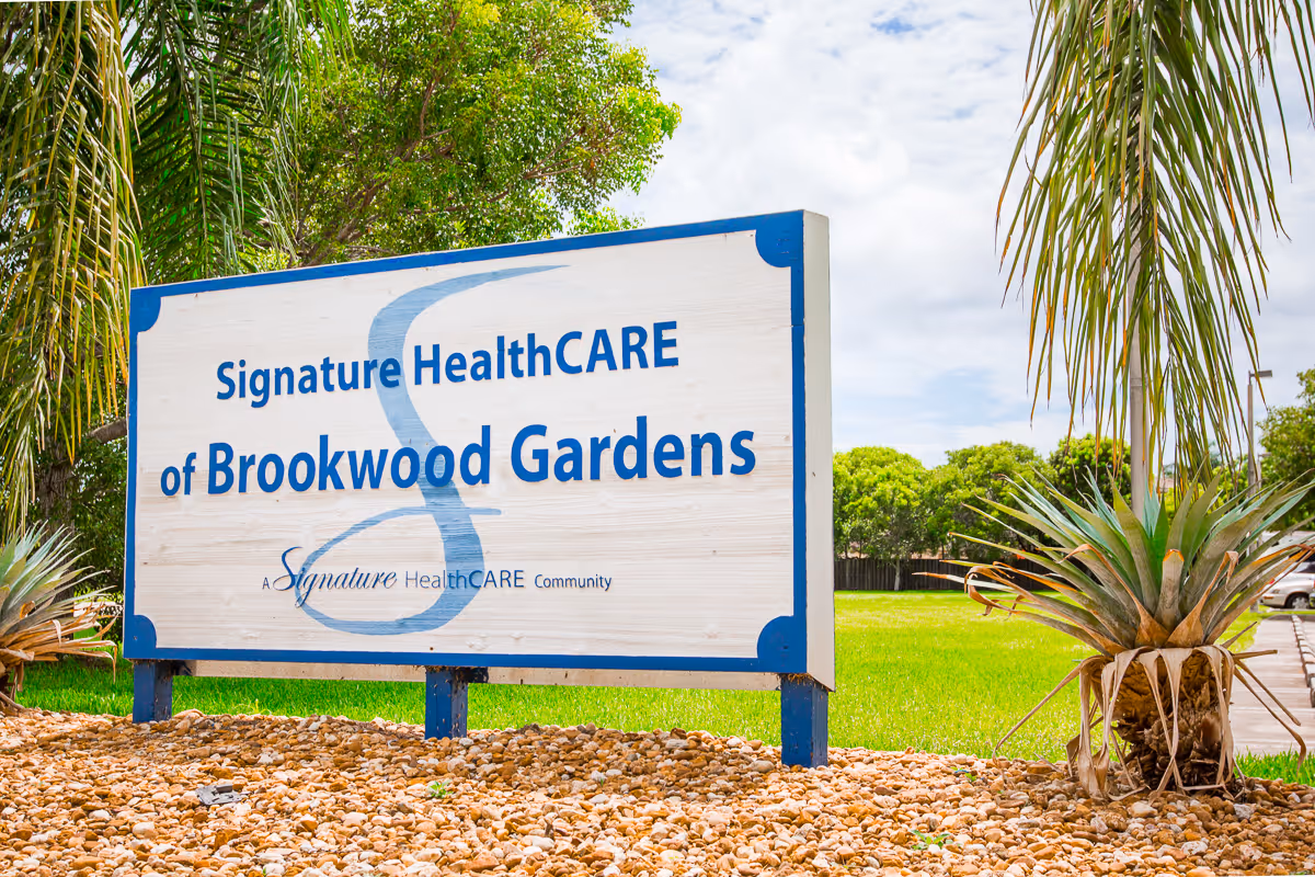 A wooden sign reading 'Signature HealthCARE of Brookwood Gardens' stands in a landscaped lawn with palm plants.