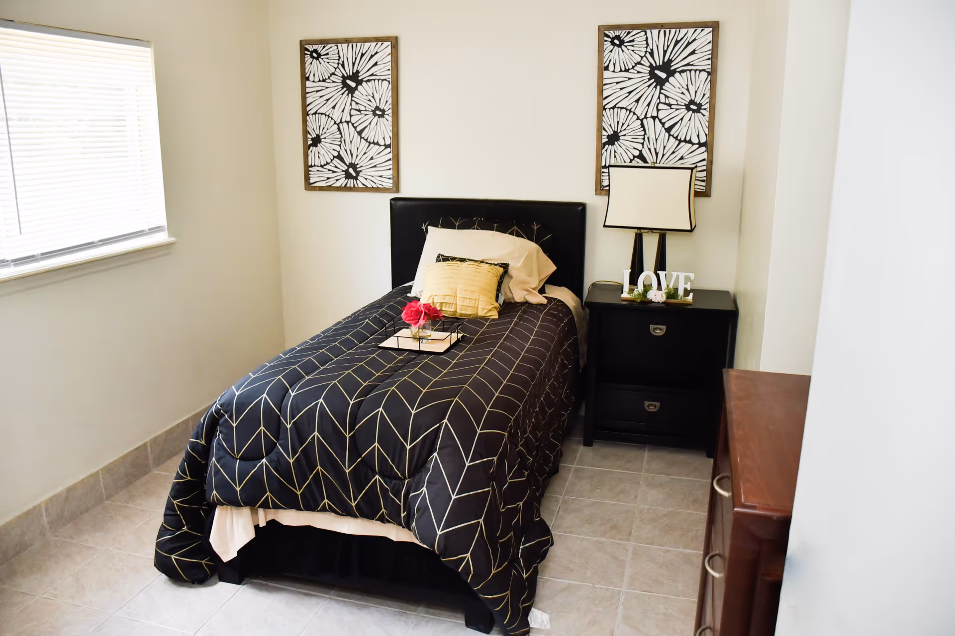 A small bedroom with a single bed covered in a black and gold patterned comforter, two pillows, and a tray with a pink flower on the bed. There is a black nightstand with a lamp and decorative letters spelling 'LOVE'. Two black and white floral artworks hang on the wall above the bed. A window with blinds is on the left side, and a wooden dresser is partially visible on the right.