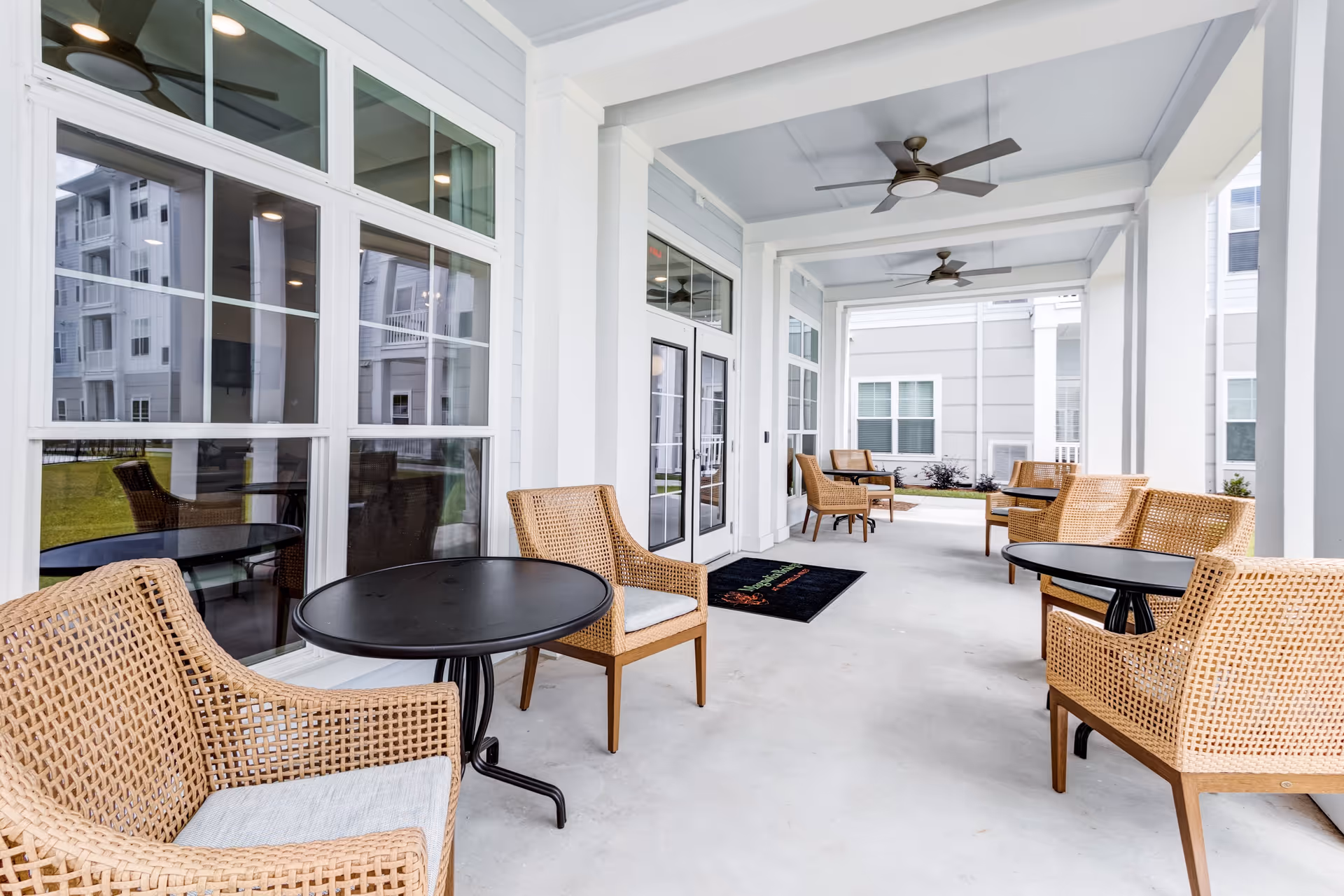 Covered outdoor seating area with multiple wicker chairs and round black tables arranged along a white building exterior with large windows and ceiling fans overhead.