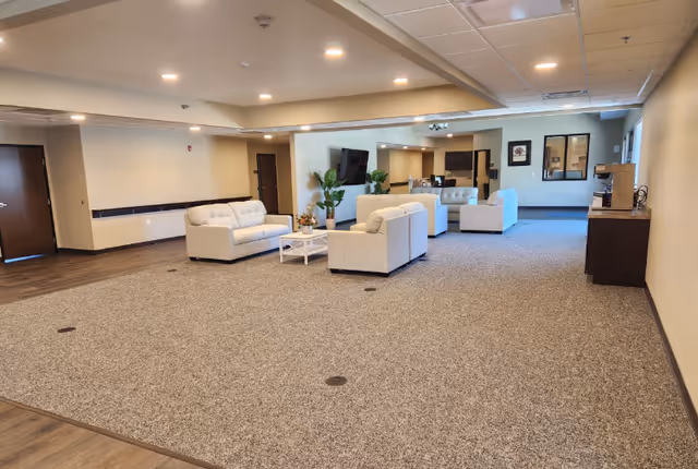 A spacious, well-lit common area with beige walls and carpeted floor. The room features several white cushioned sofas arranged around a white coffee table with a flower arrangement. There is a flat-screen TV mounted on the wall, some potted plants, and a kitchenette area with dark cabinets and a coffee machine on the right side. The ceiling has recessed lighting and a drop ceiling with tiles.
