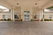 Covered entrance area of a senior living facility with double glass doors, large windows, and beige brick walls. There are potted plants and benches on either side of the entrance under a white ceiling.