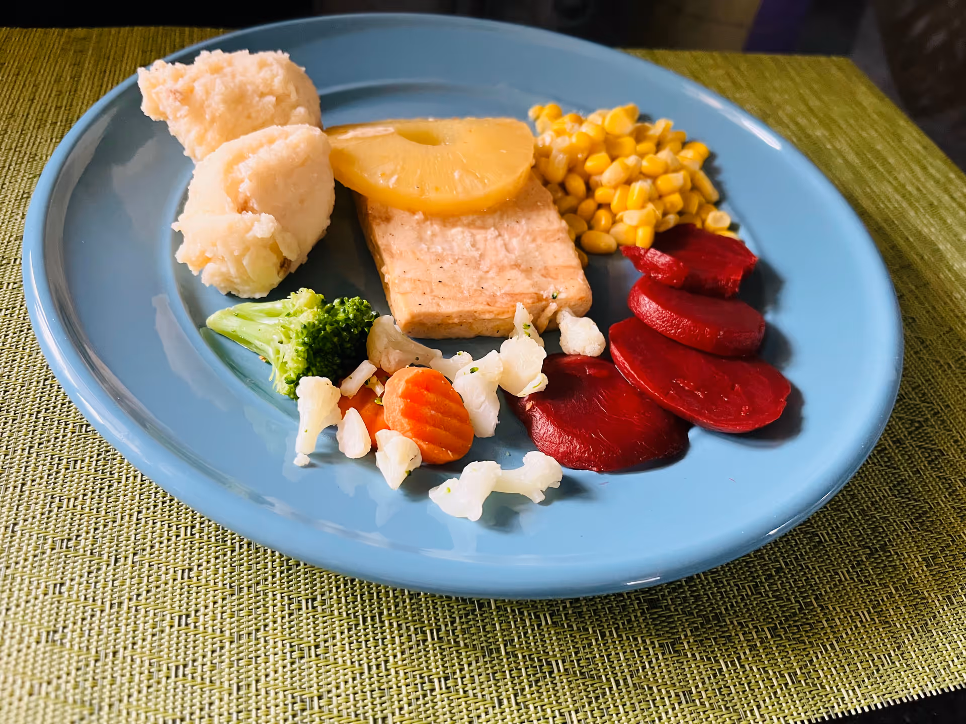 A blue plate with a serving of mashed potatoes, a slice of grilled meat topped with a pineapple slice, corn kernels, sliced beets, and a small portion of mixed steamed vegetables including broccoli, cauliflower, and carrots, placed on a green woven placemat.
